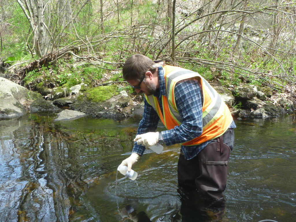 A RIDEM employee collects a water sample from a forested stream