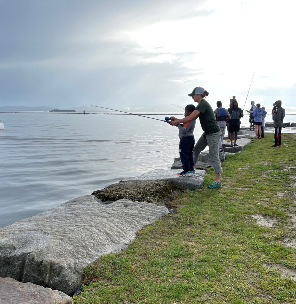 Woman teaching youth to fish along the banks of Lake Champlain.