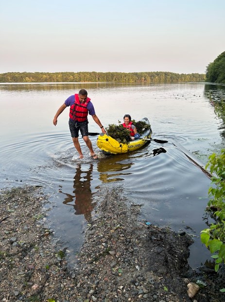 Woman in a kayak full of water chestnut plants being pulled onto the shore by a man in a red PFD.
