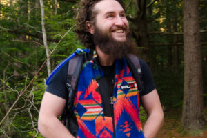 Man stands in a forest with a smile on his face and his hands in his pockets. He is wearing a blue patterned vest, grey shorts, and a hiking backpack.