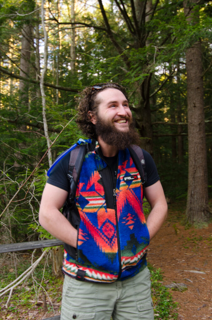 Man stands in a forest with a smile on his face and his hands in his pockets. He is wearing a blue patterned vest, grey shorts, and a hiking backpack.