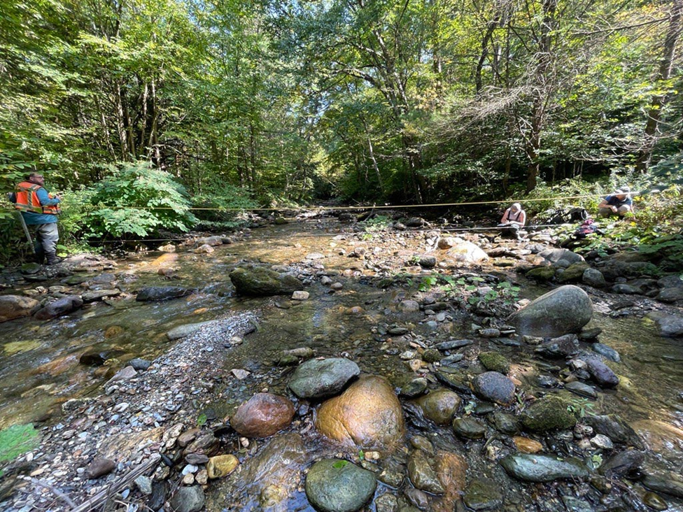 Two researchers hold two pieces of tape over the width of a stream to measure it, while another records data.