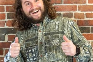 A photo of James Plummer standing in front of a brick wall smiling and giving two thumbs up.