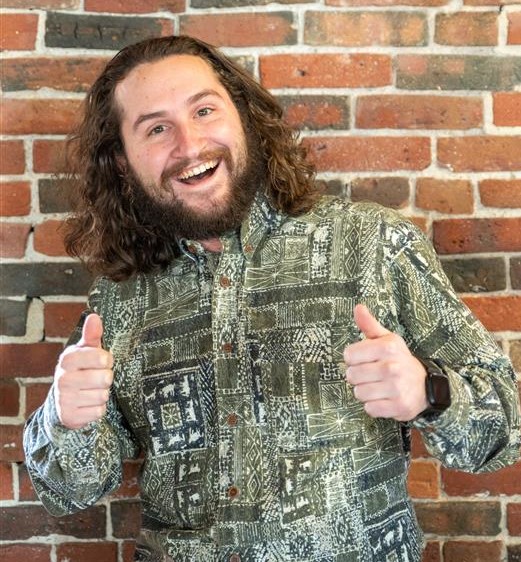 A photo of James Plummer standing in front of a brick wall smiling and giving two thumbs up.