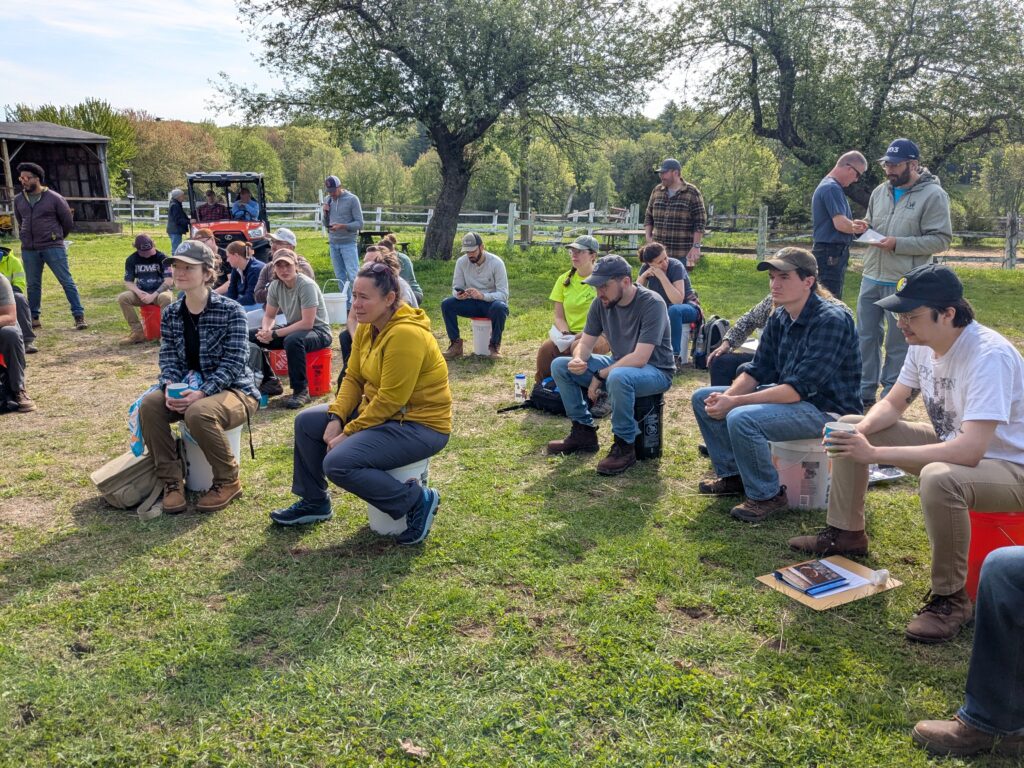 A group of students sit on buckets in a grassy field, with supplies for the class by their feet.