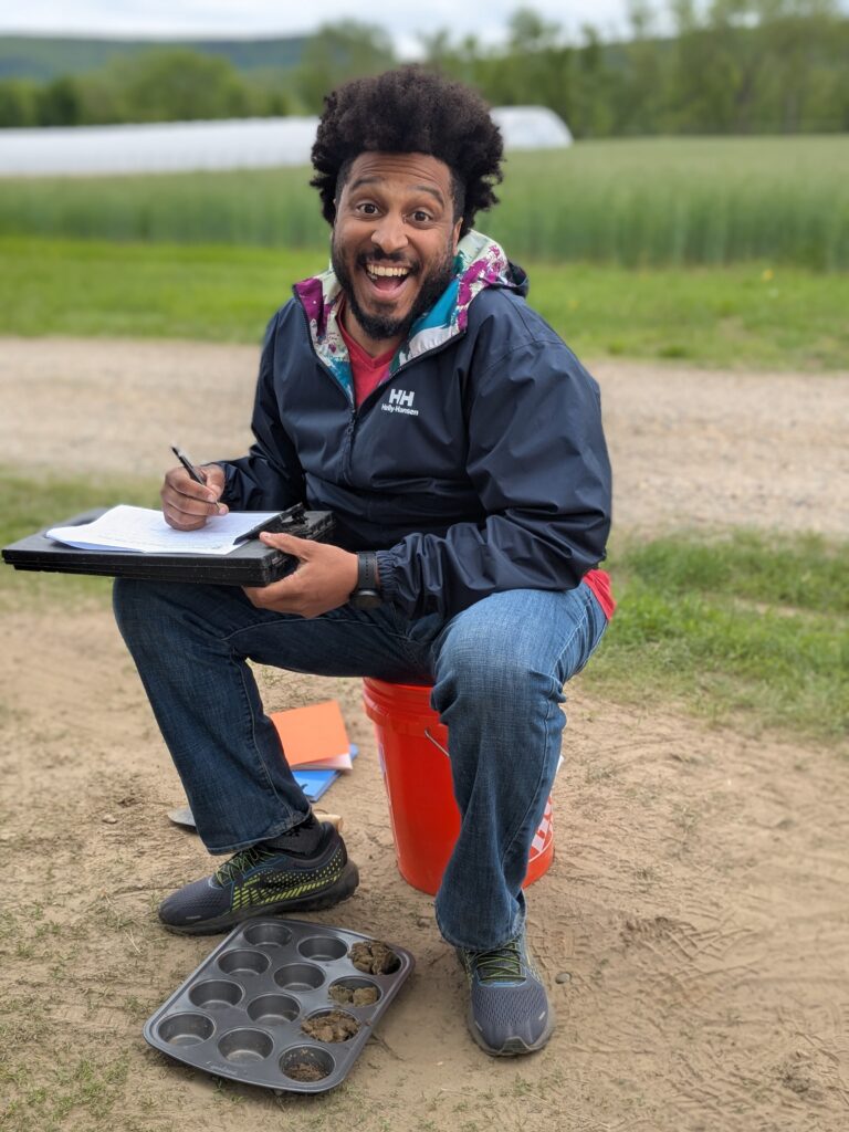 A man sits on a bucket and smiles at the camera. He has a data sheet in his sand and a cookie tray filled with soil samples by his feet.