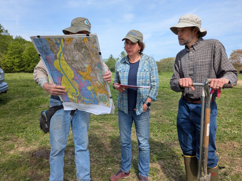 Two students looking at a topographic map held by the instructor. One student is holding a shovel. Both are dressed for the field.
