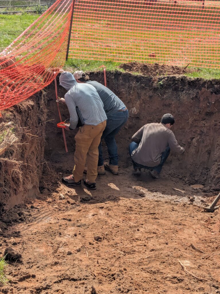 Three students squatting in a pit as they collect soil samples from the various layers.
