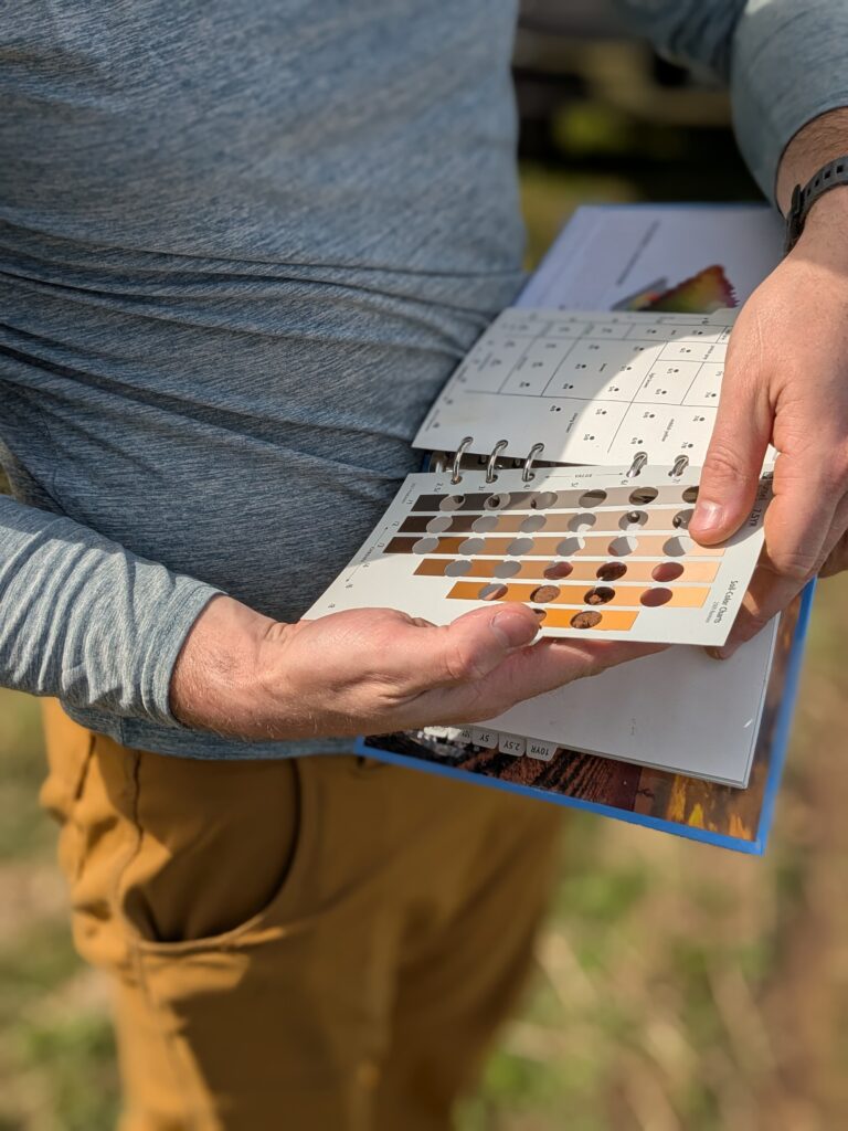 A man holding a soil color chart open. He has his hand behind the page with a soil sample as he attempts to match up the colors.