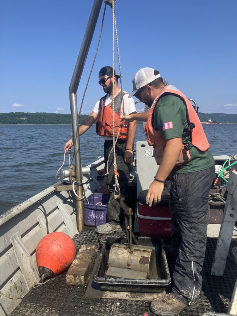 Working by boat, feld technicians use a small crane to maneuver the dredged sediment sample into a container for analysis.