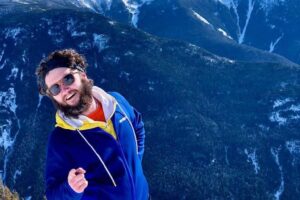 A photo of James Plummer, who is wearing a blue jacket and posing with snowy mountains in the backdrop.