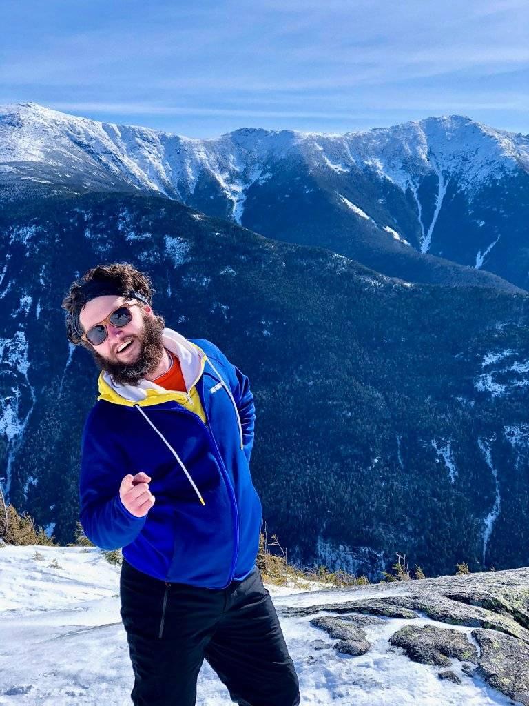 A photo of James Plummer, who is wearing a blue jacket and posing with snowy mountains in the backdrop.