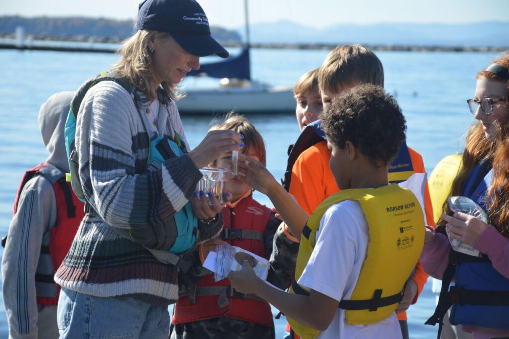 Teacher and group of youth looking at water samples in tubes while on the shore of Lake Champlain.