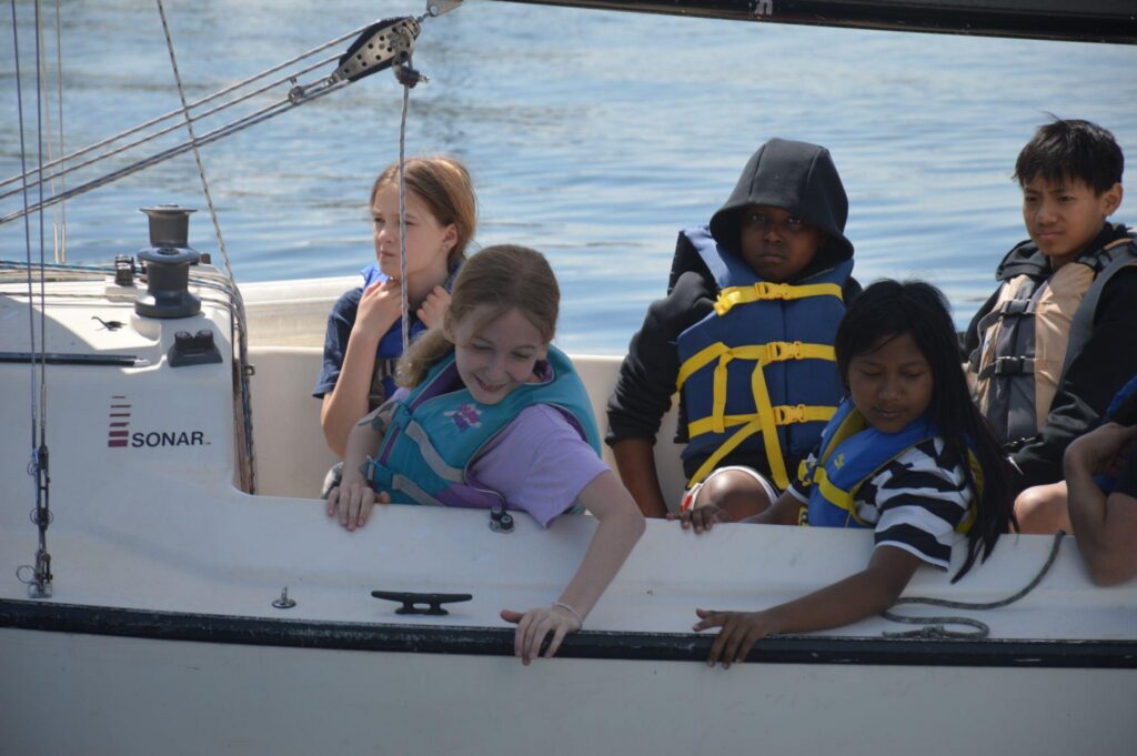 Group of kids on a sailboat wearing PFDs. Two girls looking over the side of the boat.