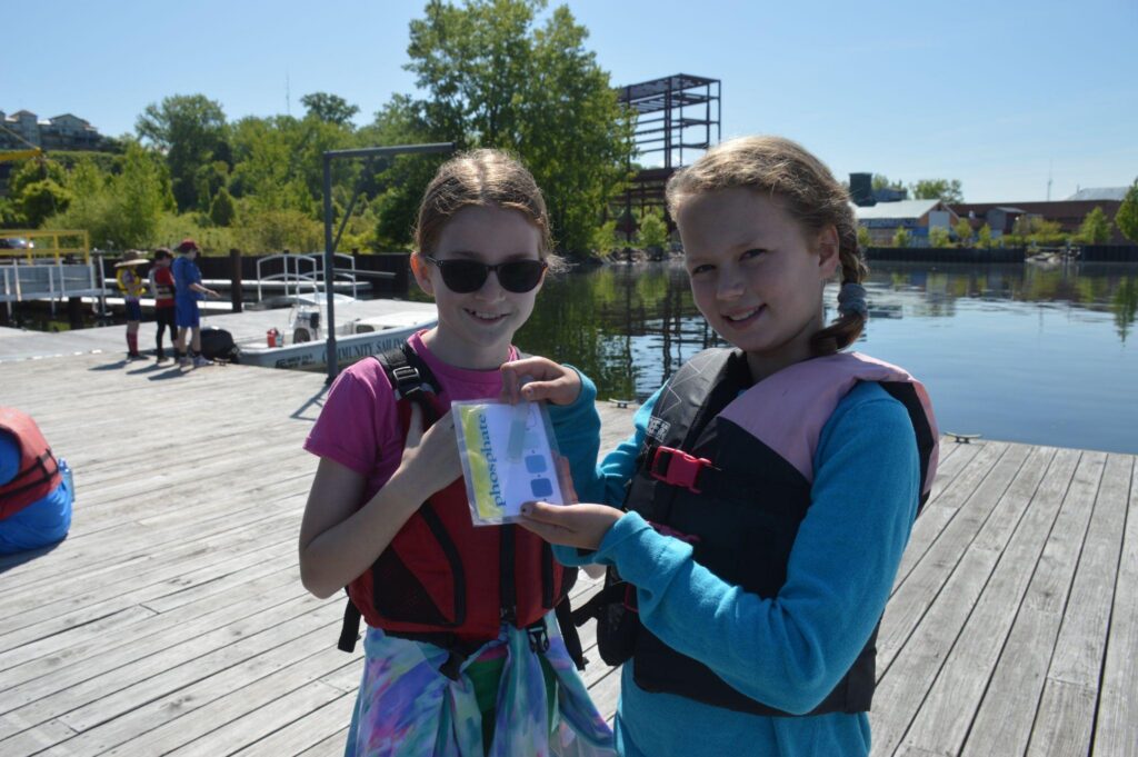 Two youth on a dock wearing PFDs post with a water sample testing kit for phophate.