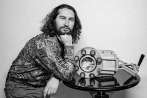 Black and white photo of James Plummer posing next to an an old tank part which is on a side table.