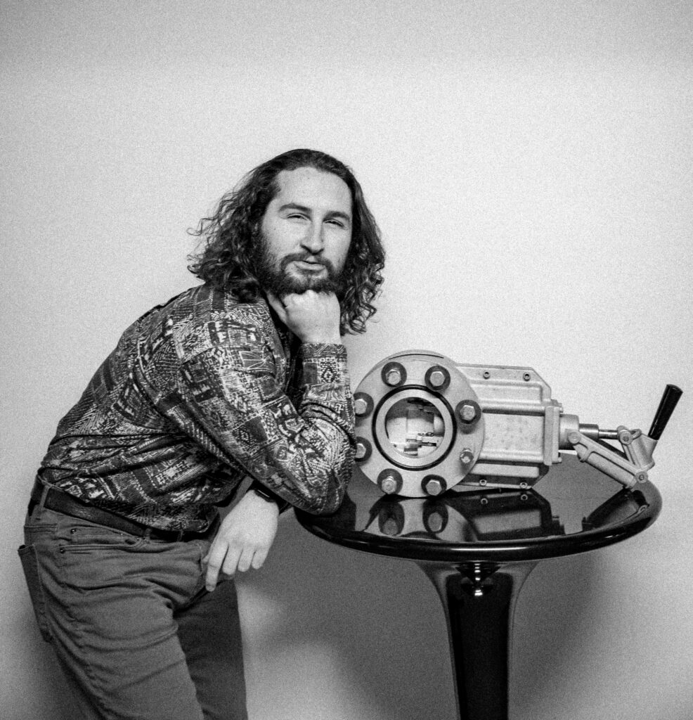 Black and white photo of James Plummer posing next to an an old tank part which is on a side table.